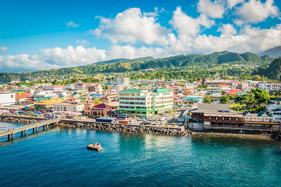 Bright, sunny view of a coastal city with colorful buildings, a harbor, and lush green mountains in the background.