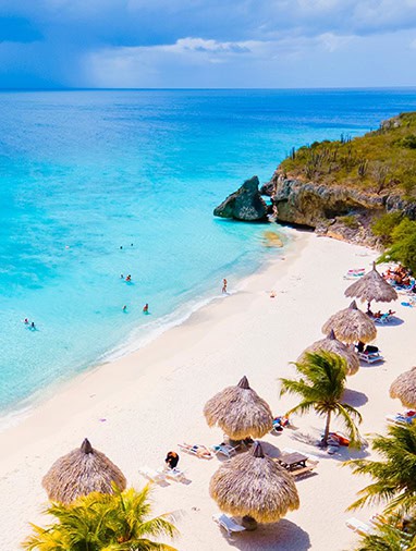 Aerial view of a white-sand tropical beach with bright blue ocean water, rocky cliffs, palm trees, and beach umbrellas