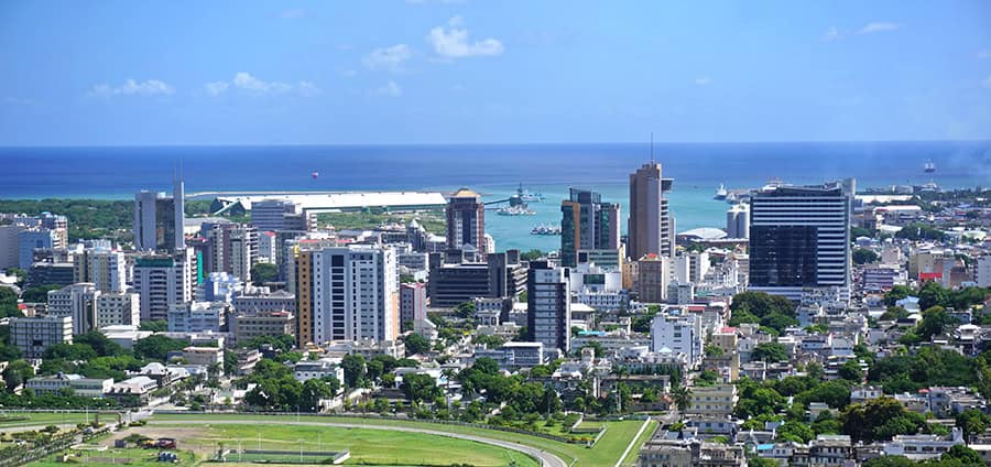 Aerial view of a coastal city skyline with tall buildings, green trees, a large harbor, and the blue ocean in the background.