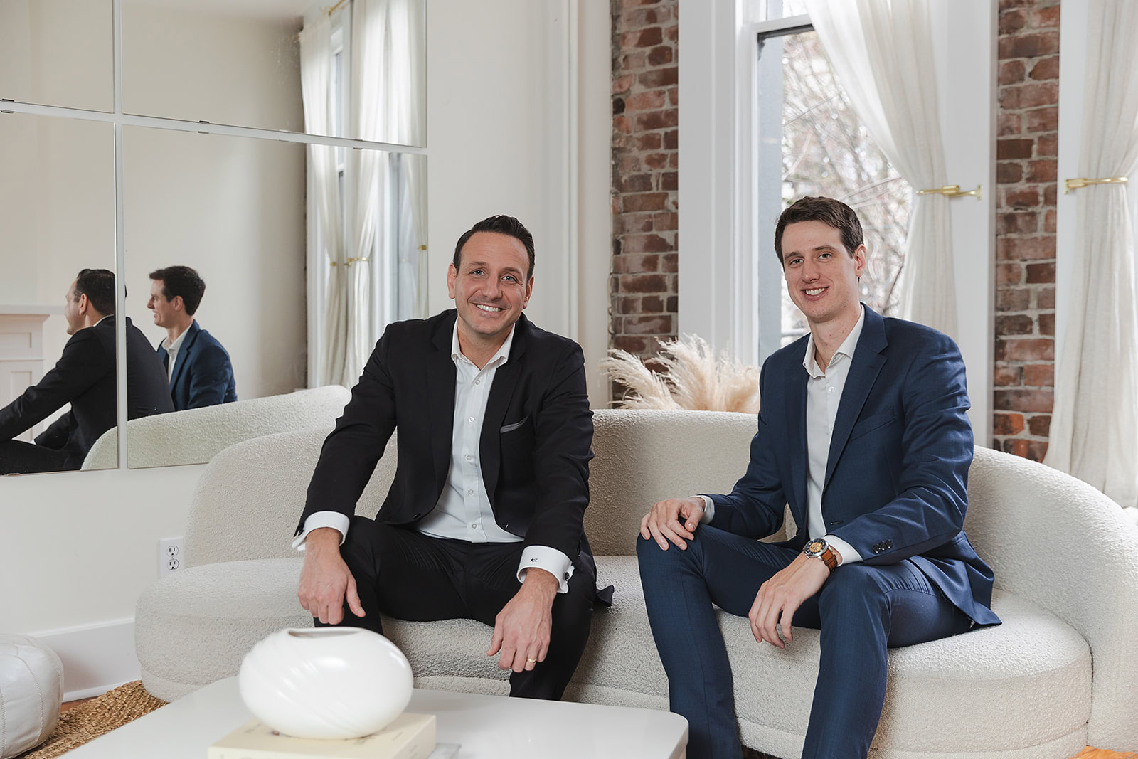 Two men in suits sit on a light-colored sofa in a modern living room, smiling at the camera. The space features brick and white walls, large windows, and a decorative vase—perfect for discussing Infinite Banking or Whole Life Insurance Policy Loans.