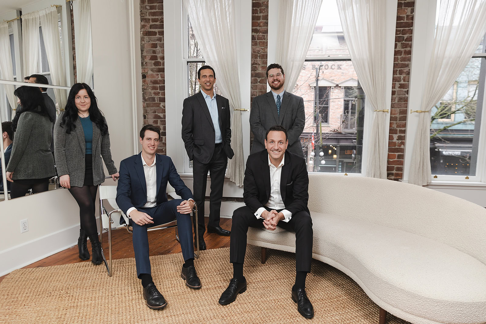 Safe Pacific Financial team professionally dressed, four men and one woman, pose in a modern office—perfect for discussing whole life insurance or infinite banking in Canada — with large windows, white curtains, exposed brick walls, a curved cream sofa, and a woven rug.