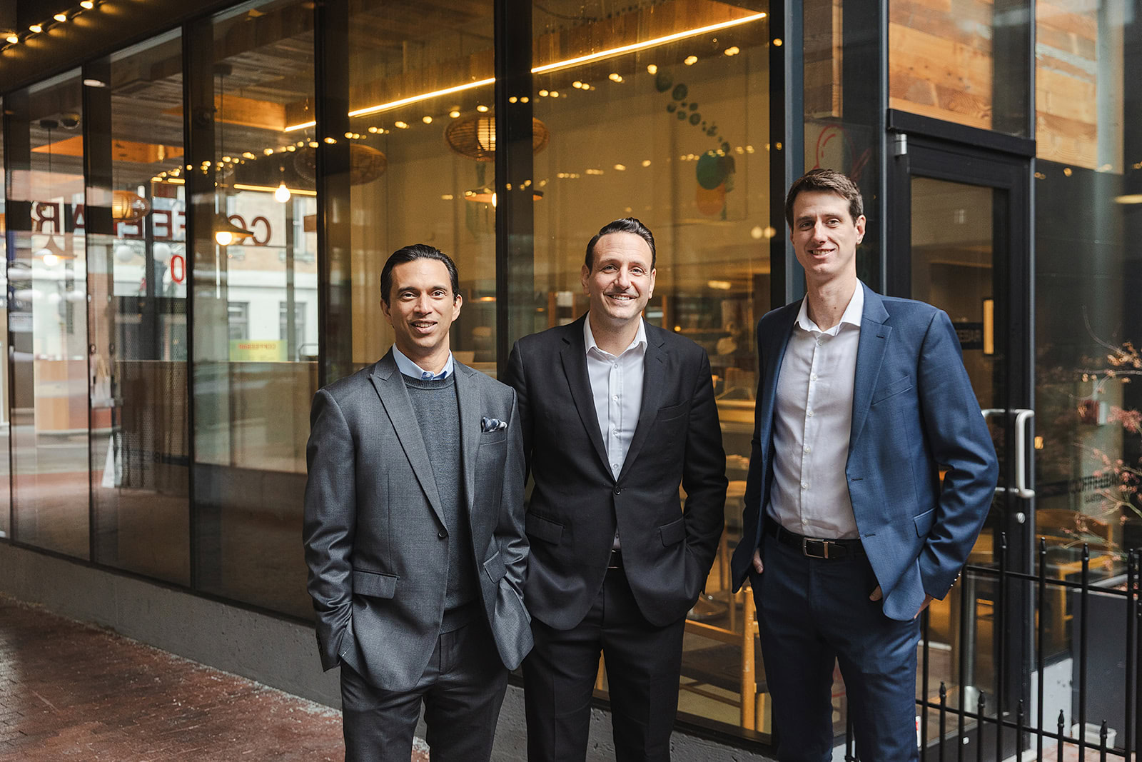 Three men wearing business suits stand together and smile in front of a modern glass building with warm lighting, suggesting a professional yet friendly atmosphere—perfect for discussing whole life insurance policy loans or infinite banking strategies.