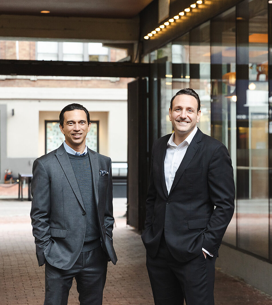 Laurent Munier and Robert Trasolini in business suits stand and smile outside a Gastown building with large glass windows, appearing confident and relaxed — perhaps discussing the Infinite Banking Concept for Canadians or Whole Life Insurance strategies on the brick-paved sidewalk.