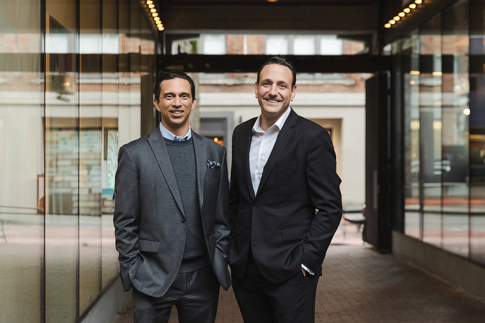 Two men wearing suits stand side by side in an outdoor walkway with glass walls, smiling with hands in their pockets. The scene, suggesting a discussion about Whole Life Insurance or Infinite Banking, features buildings visible in the background.