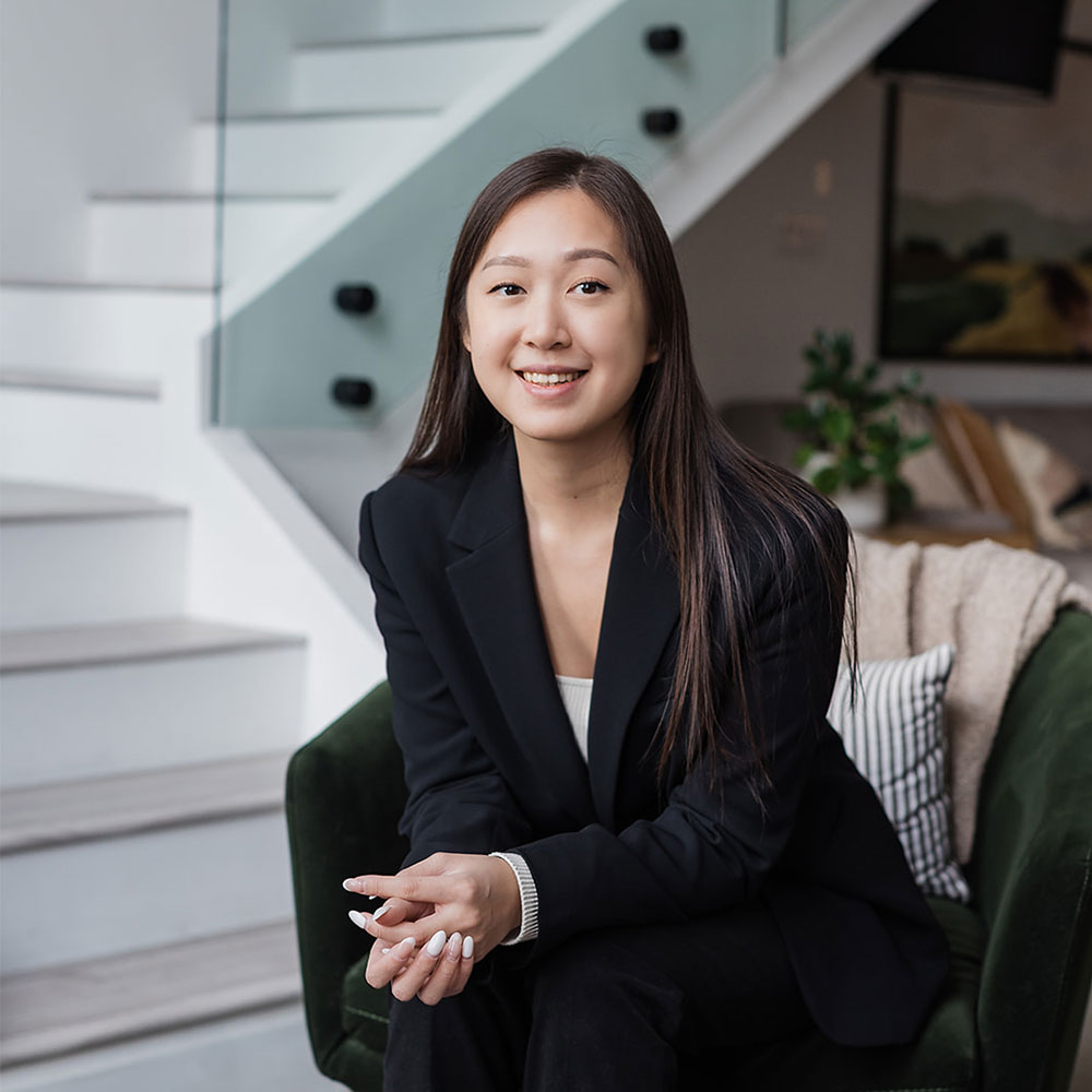 Jennifer Huang, a young woman with long brown hair, wearing a black blazer, sits and smiles in a modern living room with a staircase and green armchair in the background.