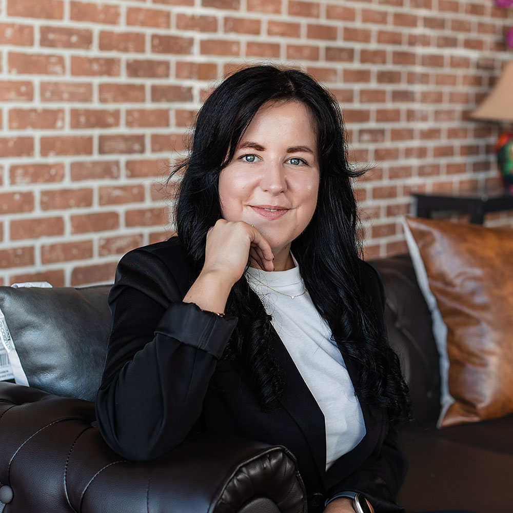 Megan Darling, a woman with long dark hair, wearing a black blazer and white shirt, sits on a brown leather couch with her hand resting on her chin. She smiles at the camera, framed by an exposed brick wall in the background.
