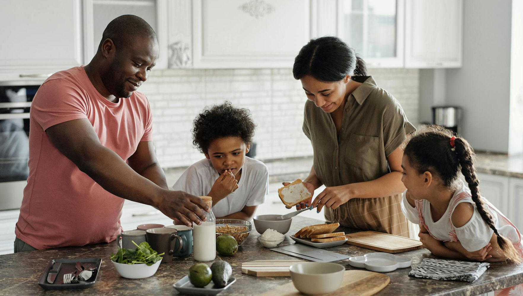 A family of four prepares food together in a bright kitchen. The parents are assembling sandwiches while their two children watch and help, all smiling and engaged in the activity.