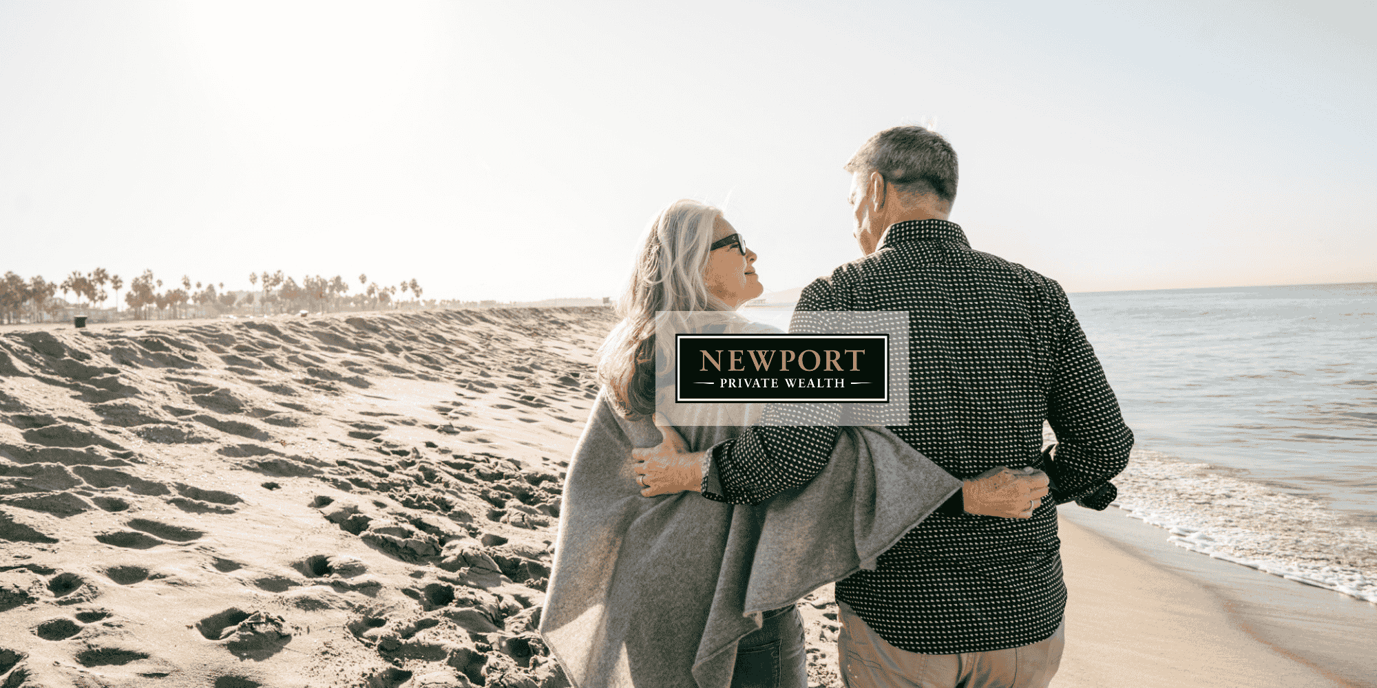 An older couple walks arm-in-arm along a sandy beach, facing the ocean—enjoying the peace of retirement made possible by thoughtful investment management in Canada. The Newport Private Wealth logo appears over the center of the image.