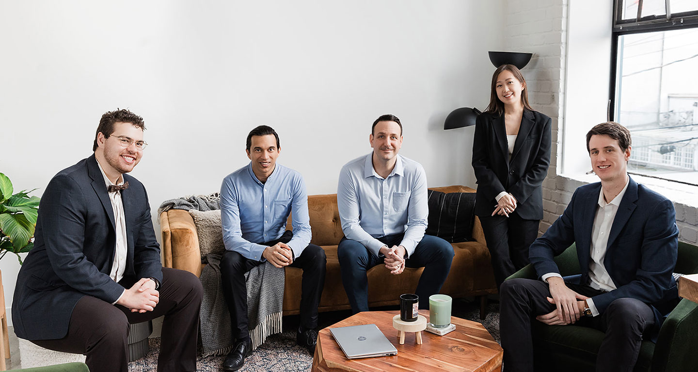 Five professionally dressed people, likely lawyers or accountants, four men sitting and one woman standing, pose together in a modern office with a sofa, armchair, coffee table, and laptop. The atmosphere is friendly and collaborative.