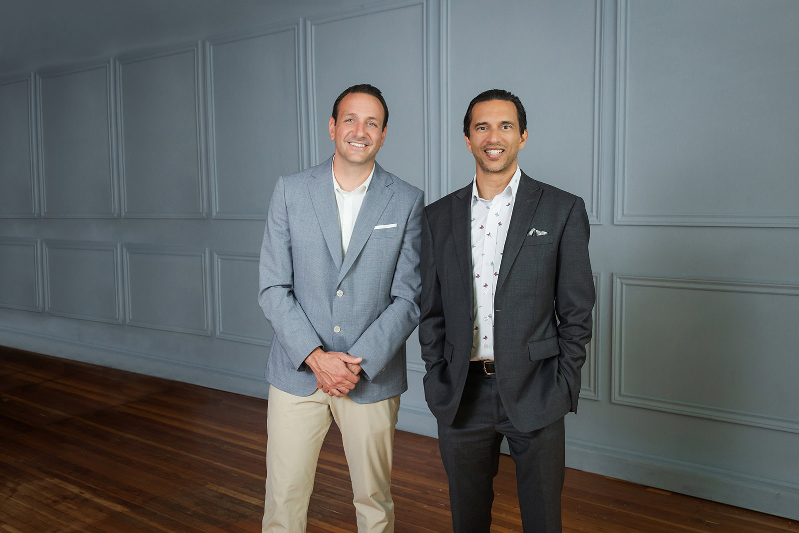 Two men stand side by side in a room with wooden floors and gray paneled walls. Both are smiling and dressed in suits; one wears a light gray jacket with khaki pants, and the other wears a dark gray suit.