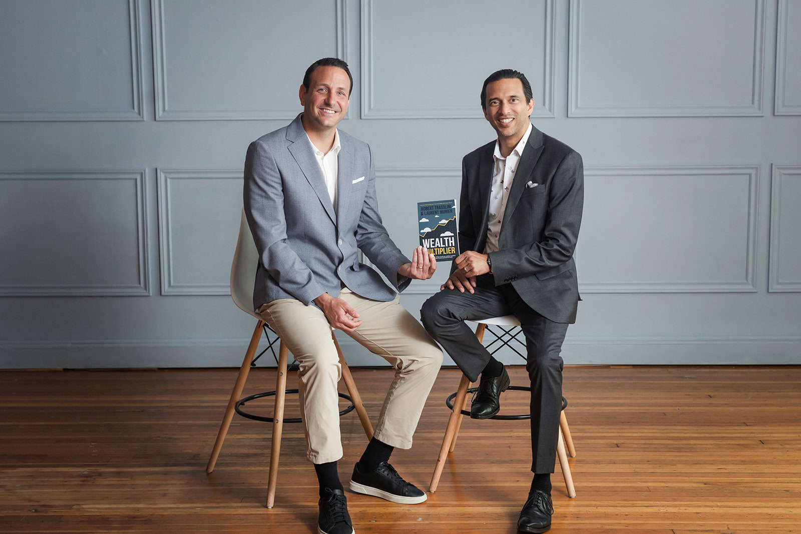 Two men in suits sit on bar stools against a gray paneled wall. One man holds a book titled Wealth Unplugged. Both are smiling and facing the camera, seated on a wooden floor.