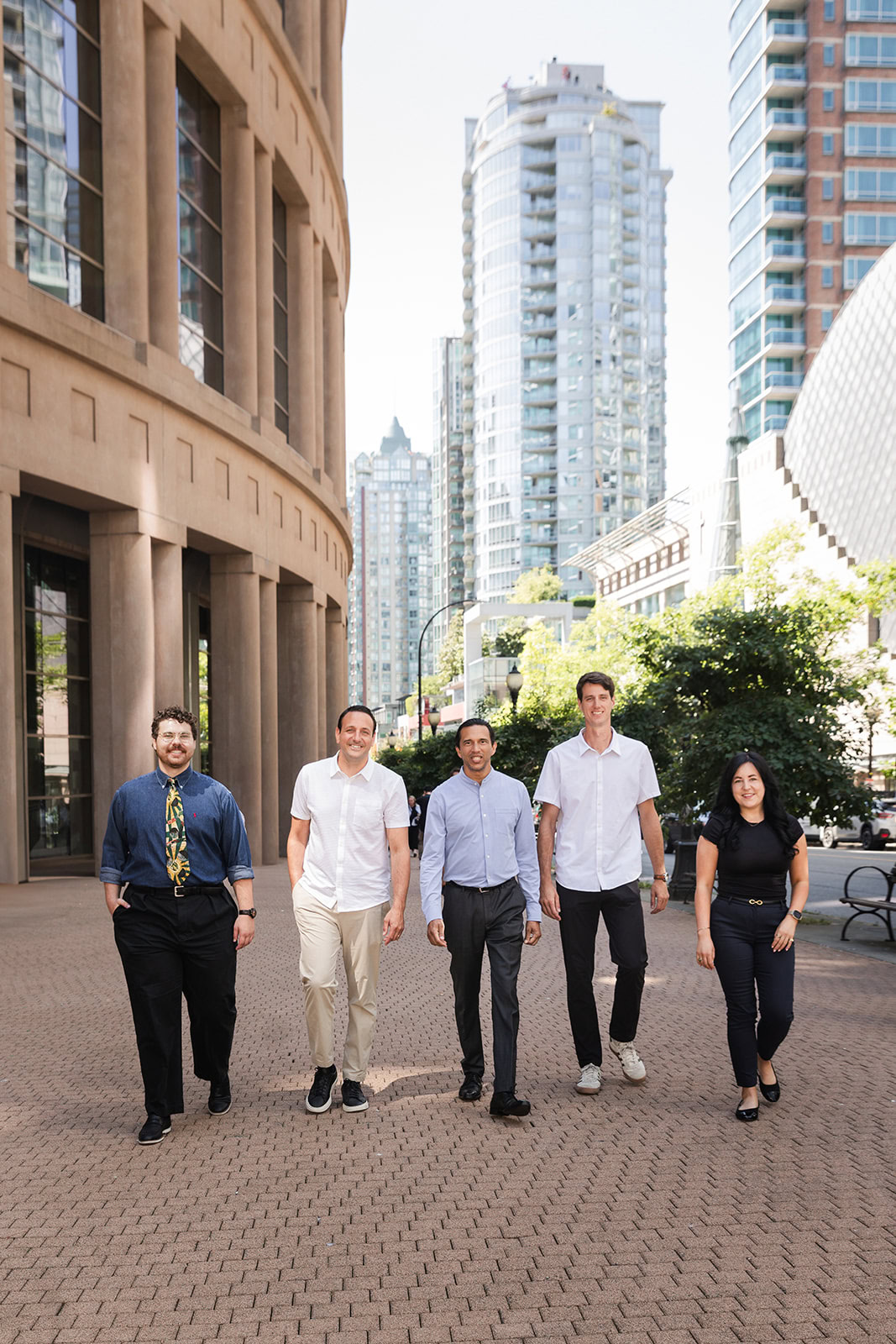 Five people walk side by side outdoors in a city, surrounded by tall buildings and greenery, on a sunny day. They are dressed in business casual attire and appear to be smiling or relaxed.