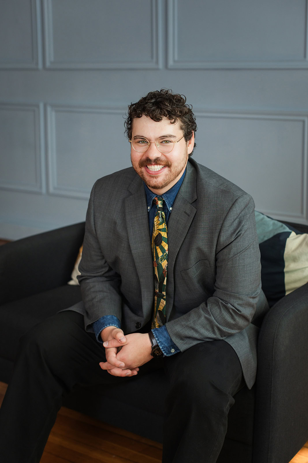 A man with curly hair, glasses, and a beard sits on a dark sofa, smiling at the camera. He wears a gray suit, a patterned tie, and a blue shirt, with his hands clasped in front of him.