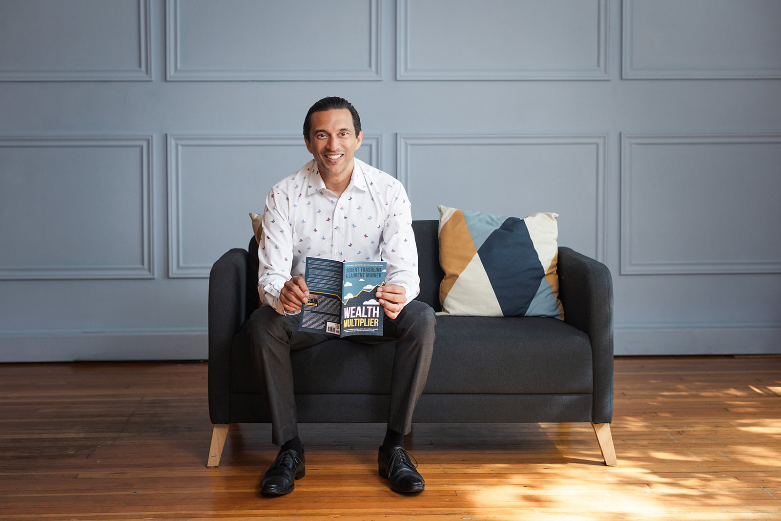 A man in a white shirt and dark pants sits on a black sofa with two patterned cushions, smiling and holding an open book titled Wealth Multiplier. The background is a light grey paneled wall and wooden floor.