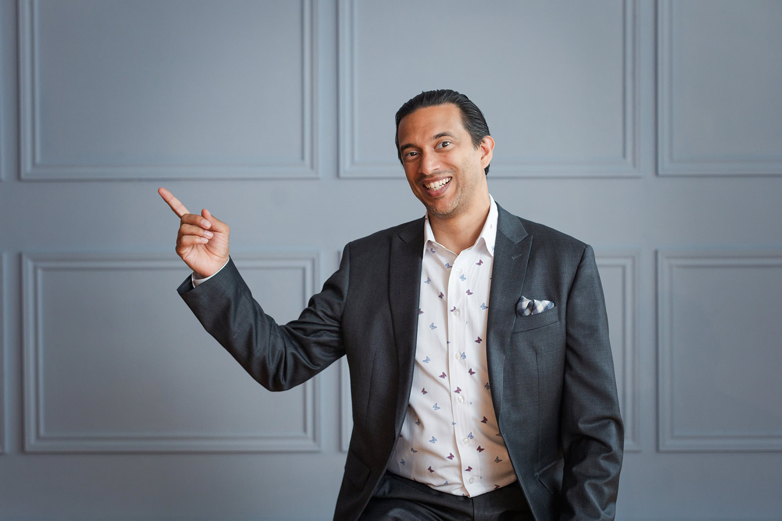 A man in a dark suit and patterned white shirt smiles while sitting and pointing to his left. He is in front of a gray wall with rectangular panel details.