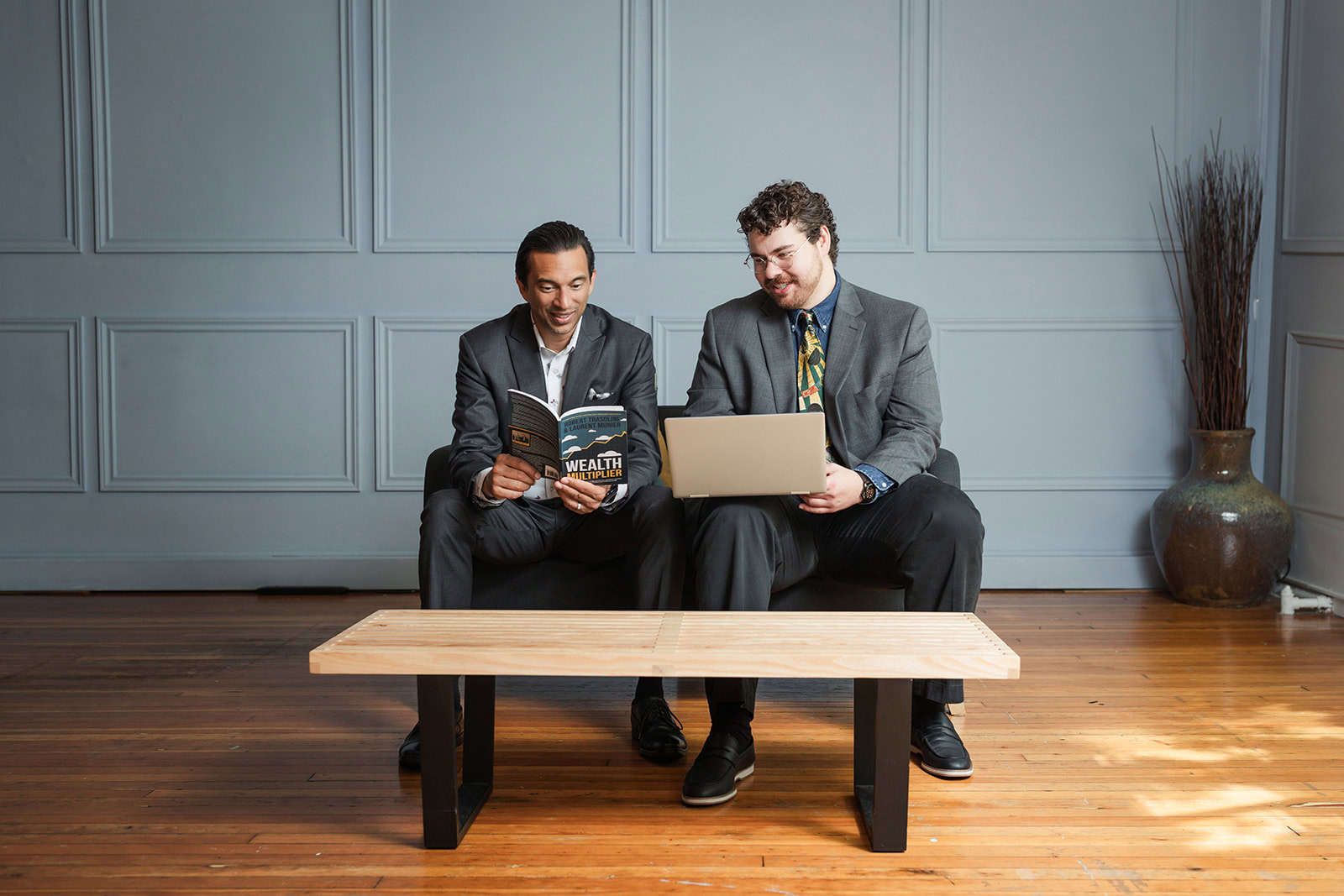 Two men in suits sit on a bench against a gray paneled wall; one reads a book titled Wealth, while the other uses a laptop. A large ceramic vase with tall branches stands to their right.