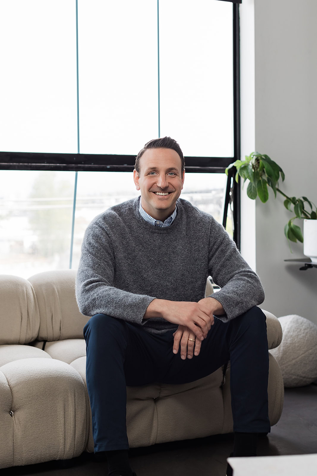 A man in a gray sweater and blue pants sits smiling on a beige sofa in a modern, bright room with large windows and a potted plant in the background.