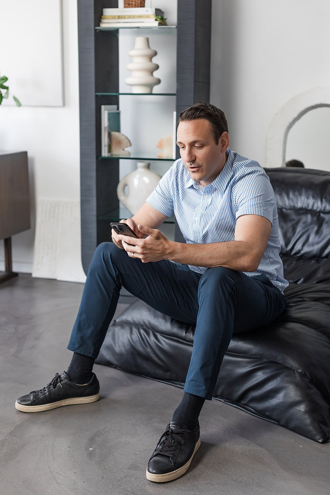 Robert Trasolini financial advisor in a short-sleeve, striped button-up shirt and dark pants sits on a black couch, looking at his phone. He is indoors, with shelves and decorative objects visible in the background.