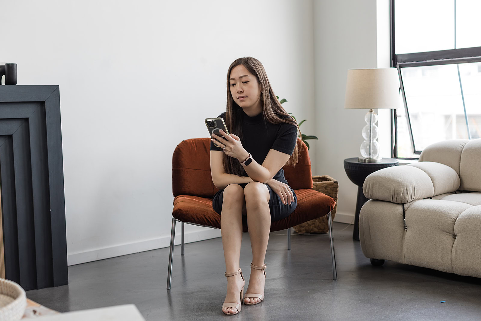 A woman sits on a rust-colored chair in a modern living room, looking at her phone. She is dressed in a black outfit and heels. The room has a white sofa, a lamp, and large windows letting in sunlight.
