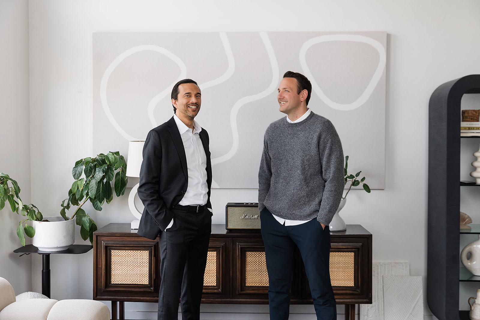 Laurent Munier and Robert Trasolini stand indoors, smiling at each other. They are discussing infinite banking in Canada is it right for you? One wears a black suit and white shirt; the other wears a gray sweater and blue pants. Abstract art and plants decorate the modern, bright room behind them.