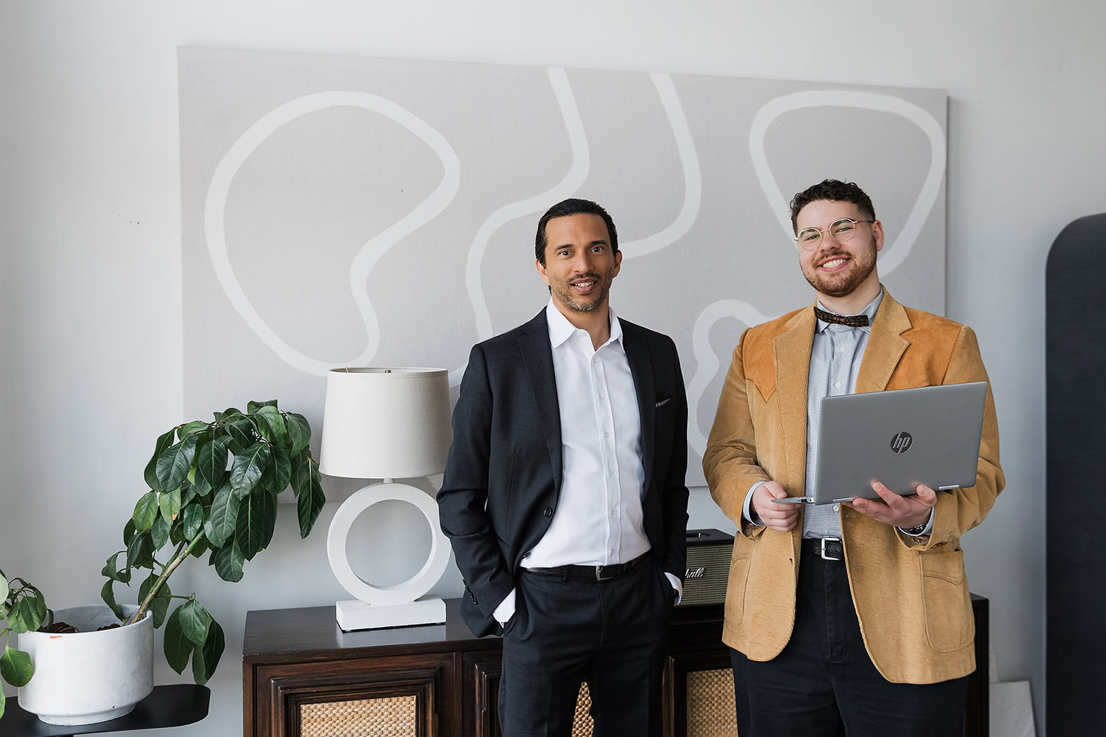Two men stand indoors in front of abstract wall art. One wears a black suit with a white shirt, the other wears glasses, a brown jacket, and holds an HP laptop. A plant and lamp sit on a cabinet beside them.