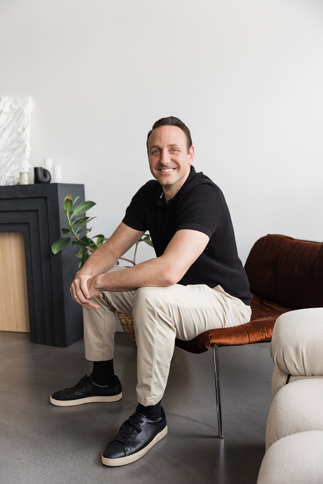 Robert Trasolini financial advisor with short brown hair, wearing a black shirt, beige pants, and black sneakers, sits on a brown chair in a modern, minimal living room with plants and neutral decor.