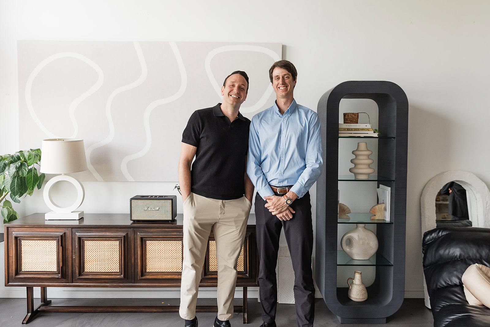 Two men stand side by side, smiling, in a modern living room with minimalist decor, including a cabinet, lamp, abstract wall art, and a glass display shelf with decorative objects.