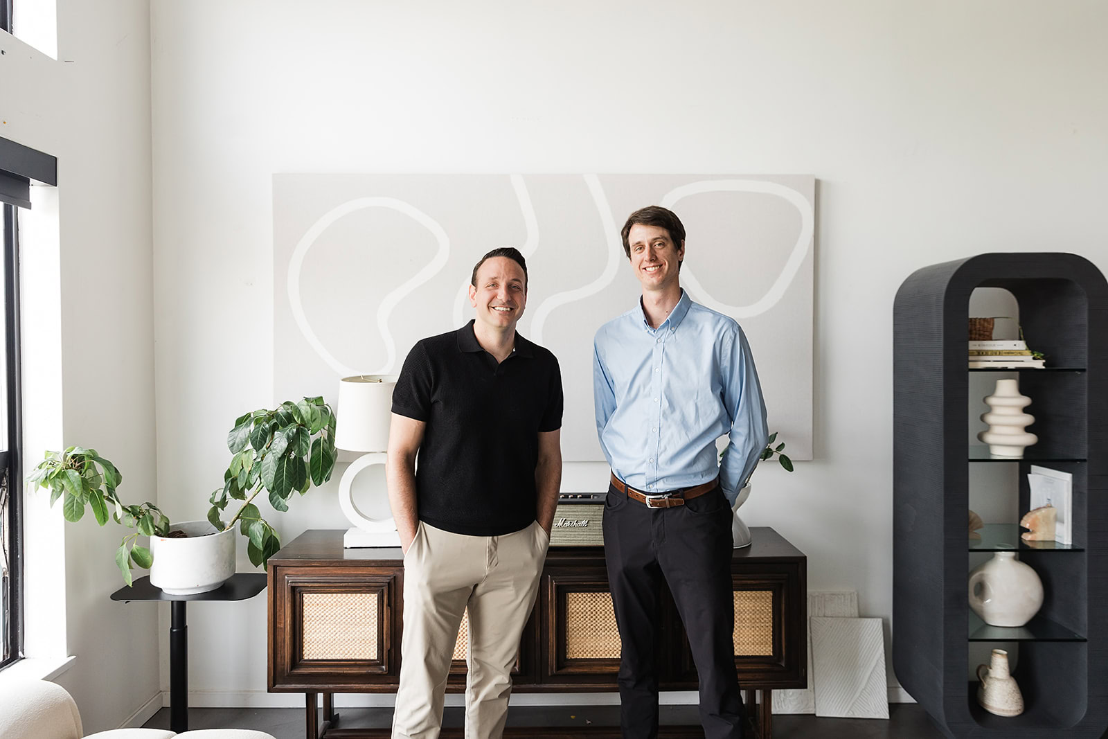 Two men stand smiling in a modern, stylish living room with minimalistic decor, plants, and abstract art on the wall behind them. One wears a black shirt and beige pants, the other a light blue shirt and dark pants.
