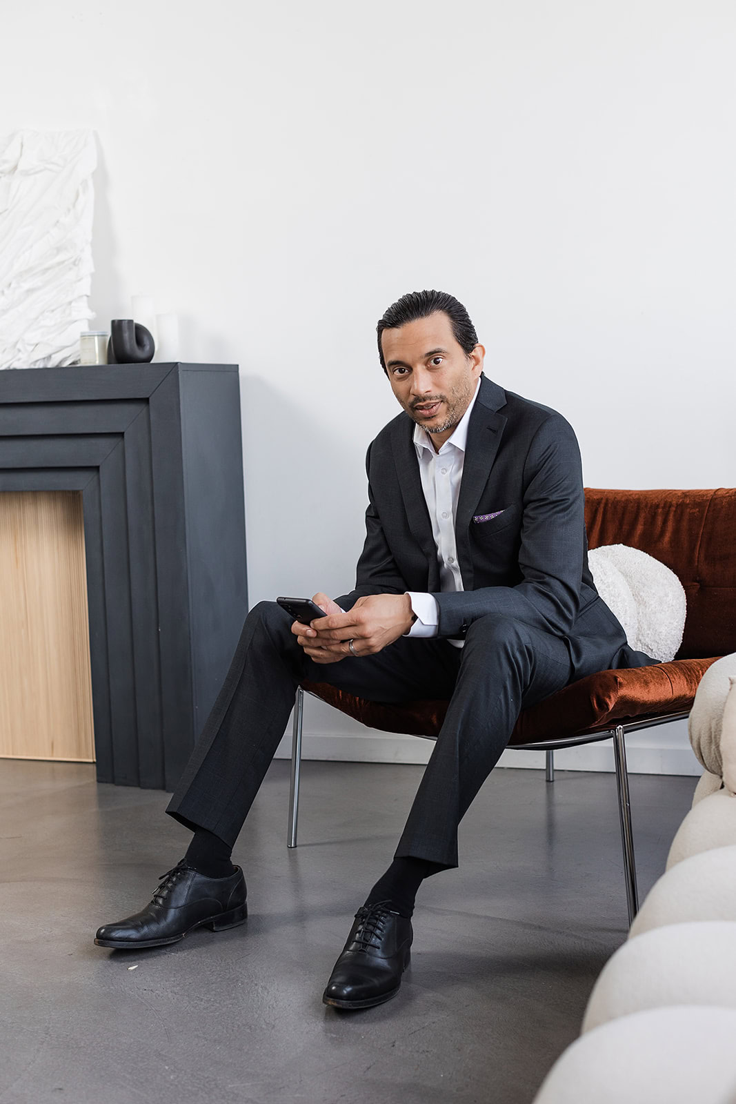 A man in a dark suit sits on a brown chair, holding a smartphone and looking forward. He is indoors, near a modern black fireplace with decorative objects, and the setting appears to be an office or lounge.