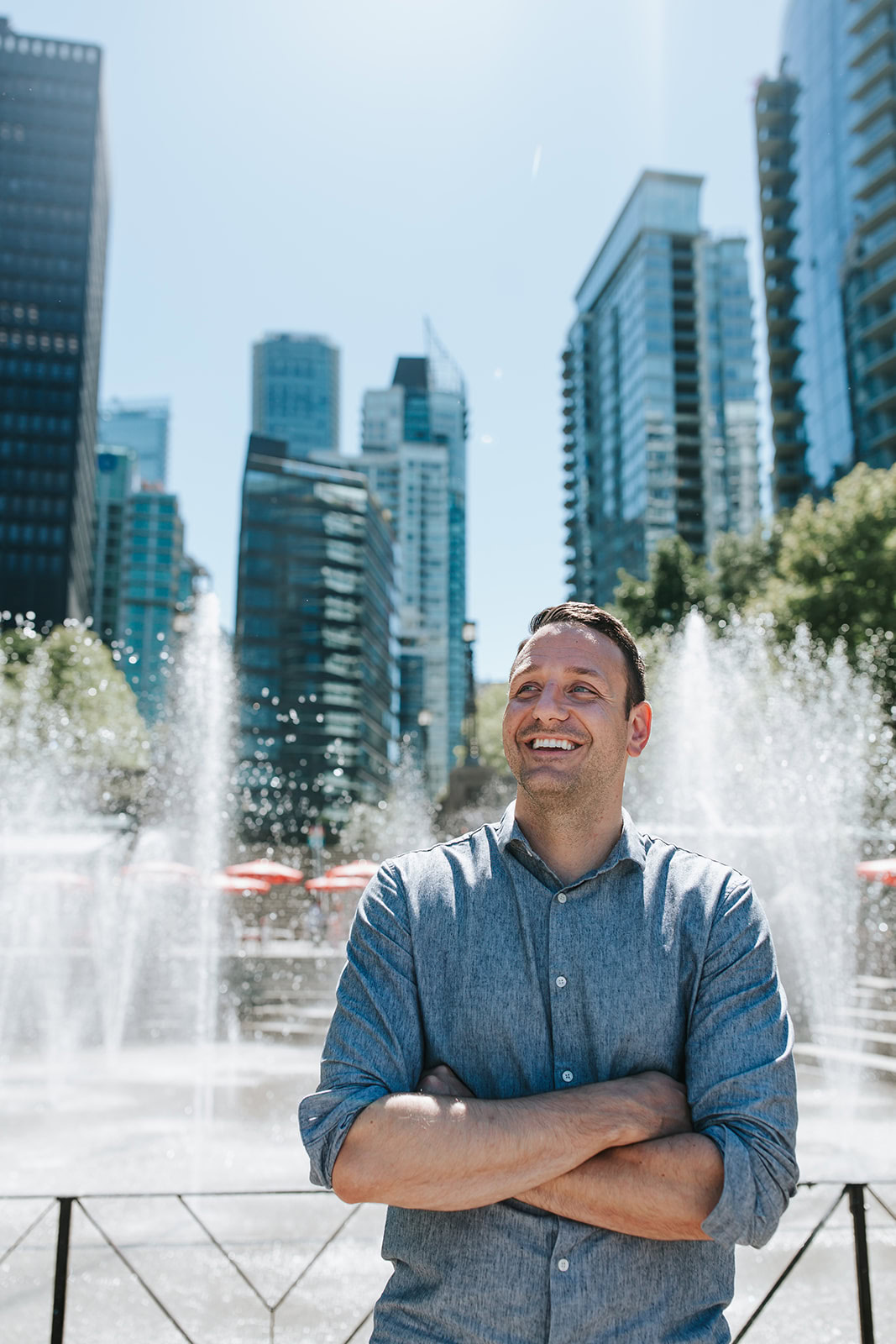 A man stands smiling with arms crossed in front of a fountain, with tall city buildings and trees in the background on a sunny day.