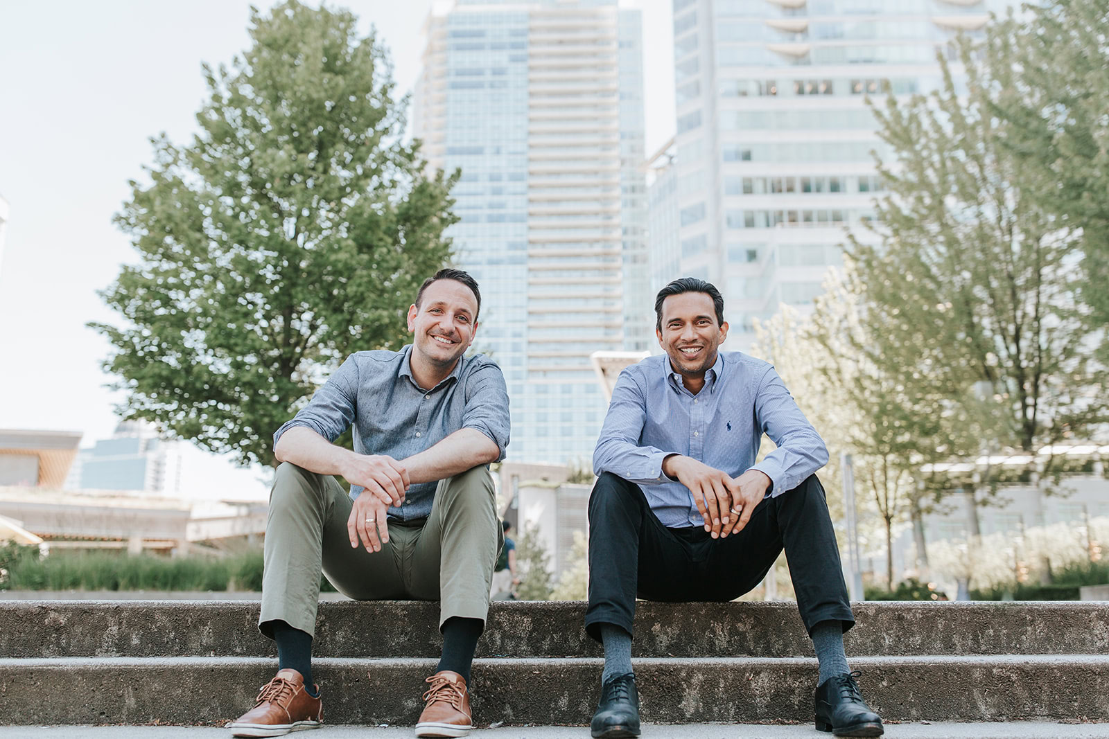 Two men sit on outdoor steps, smiling at the camera. Behind them are trees and tall modern buildings on a sunny day. Both are casually dressed in button-up shirts and trousers.
