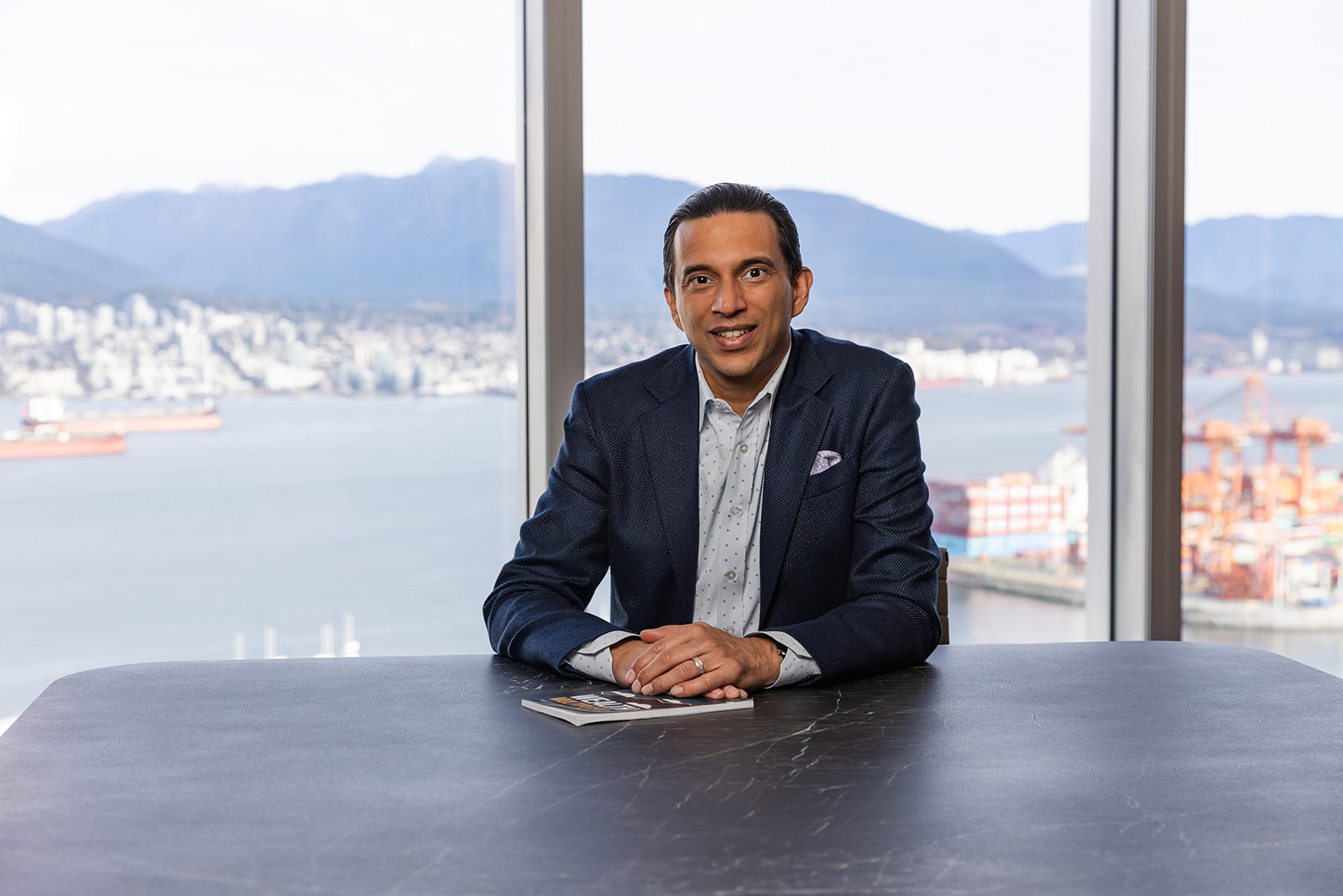 A man in a navy blazer smiles at a table with Safe Pacific Financial magazines, overlooking a cityscape—bespoke wealth management advice.