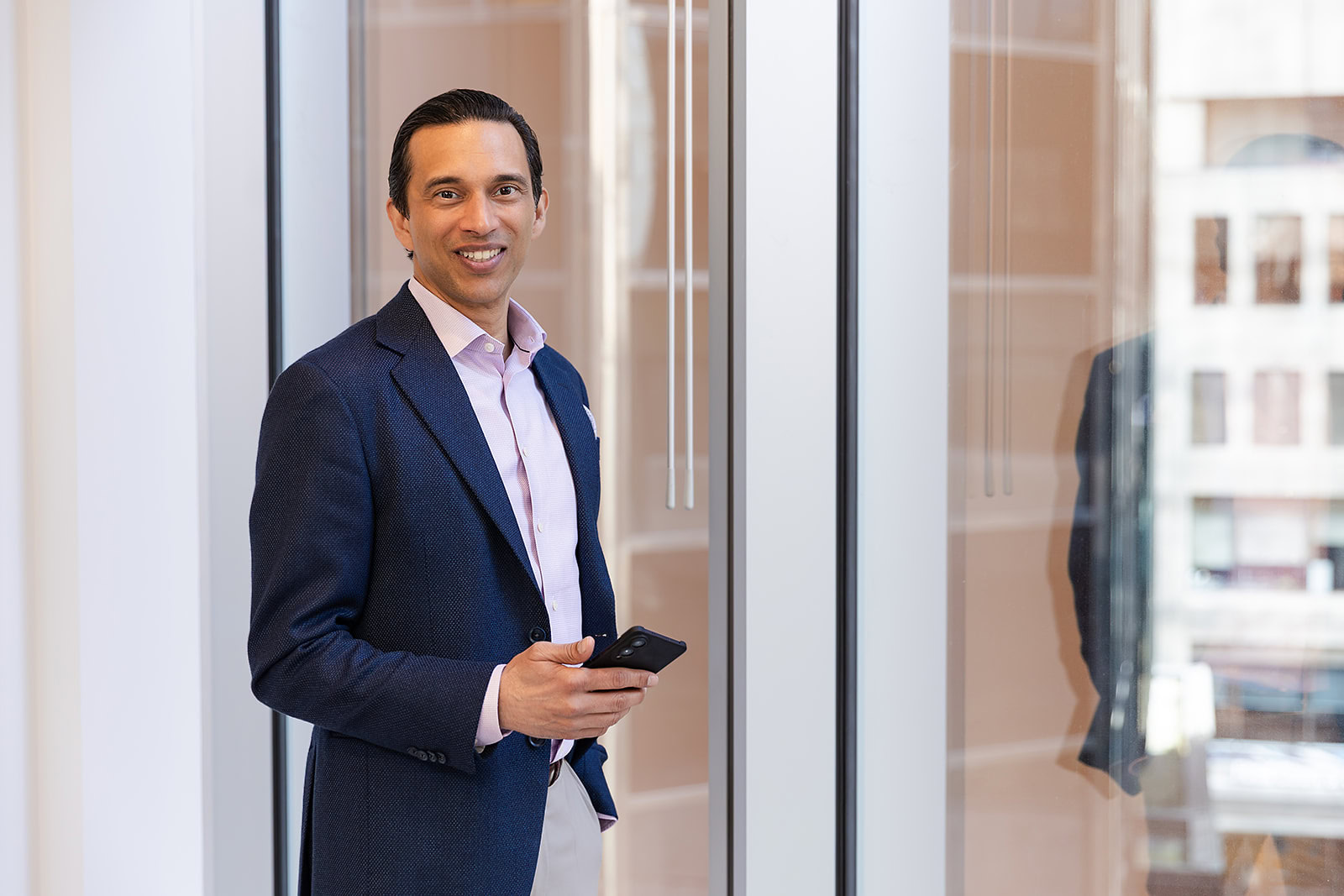 A man in a blue blazer holds a phone by a window, city view reflected—highlighting Safe Pacific’s bespoke Canadian wealth management and infinite banking strategies.