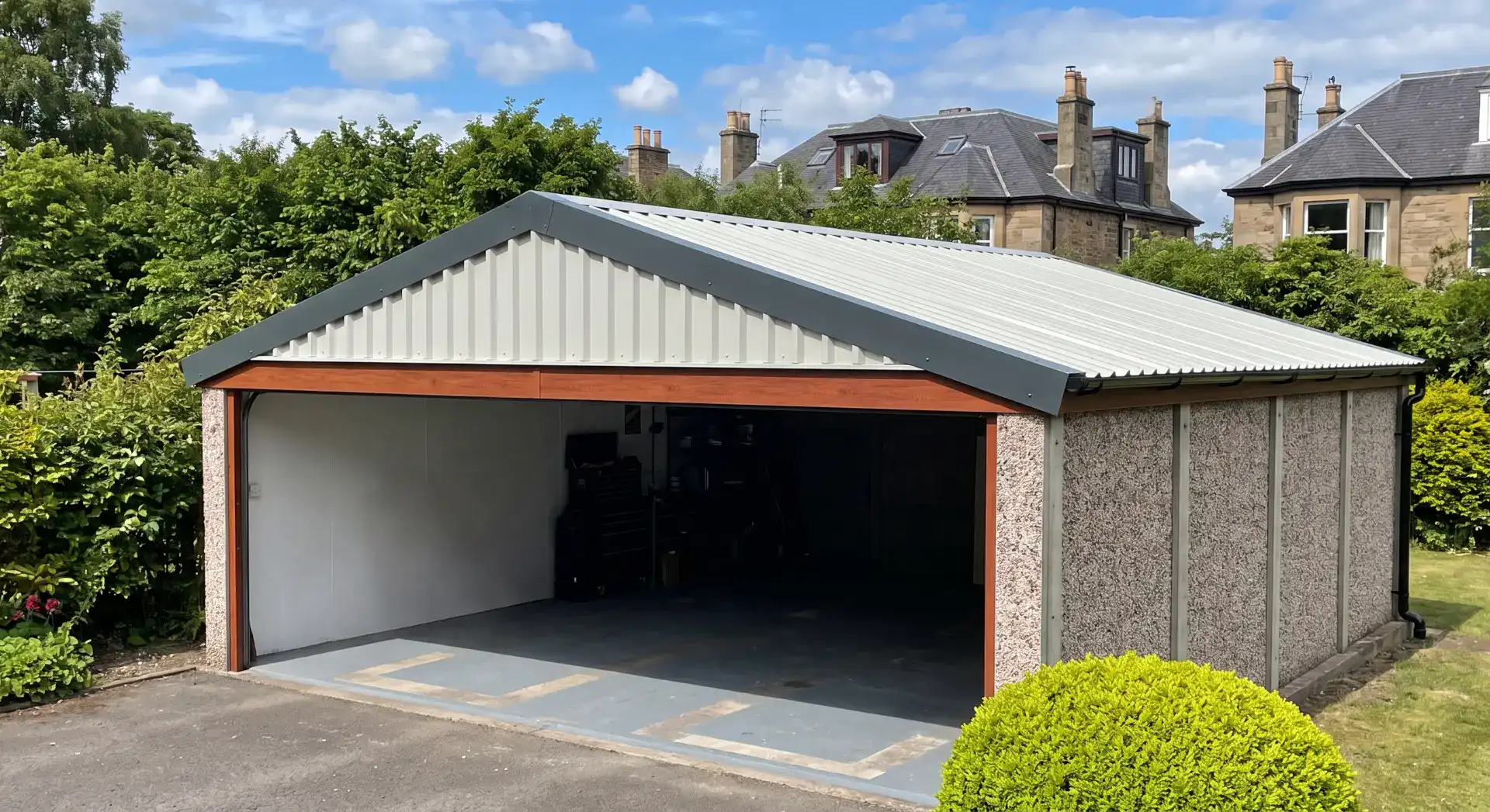 Photograph of a refurbished concrete panel garage with a newly installed clean light grey metal profile roof, dark fascia, and timber trim, set within a well-maintained garden.