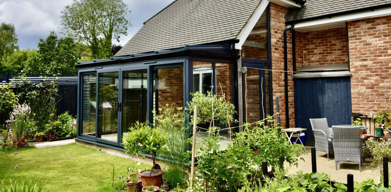 Dark framed combination conservatory with brick walls, full-height glazing and garden patio seating.