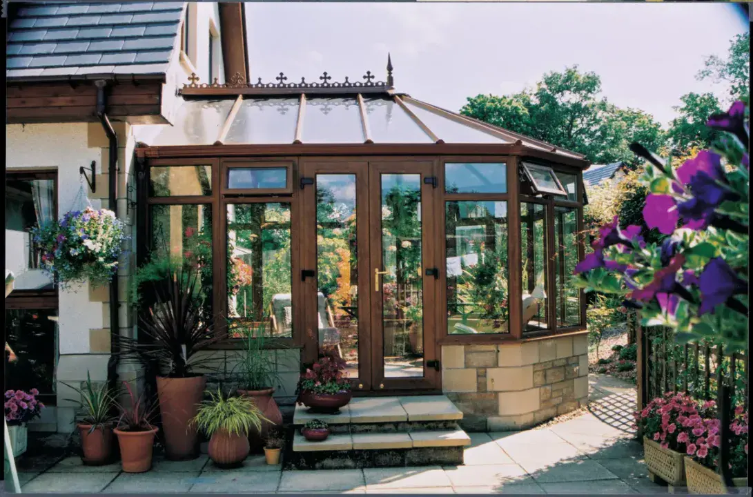 Brown Victorian conservatory with glazed roof, double doors and stone base surrounded by garden plants.