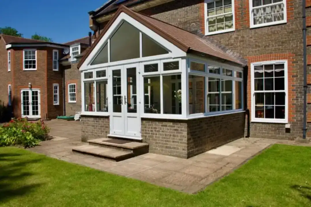 Gable end conservatory with white frames, pitched tiled roof and double doors leading to a garden patio.