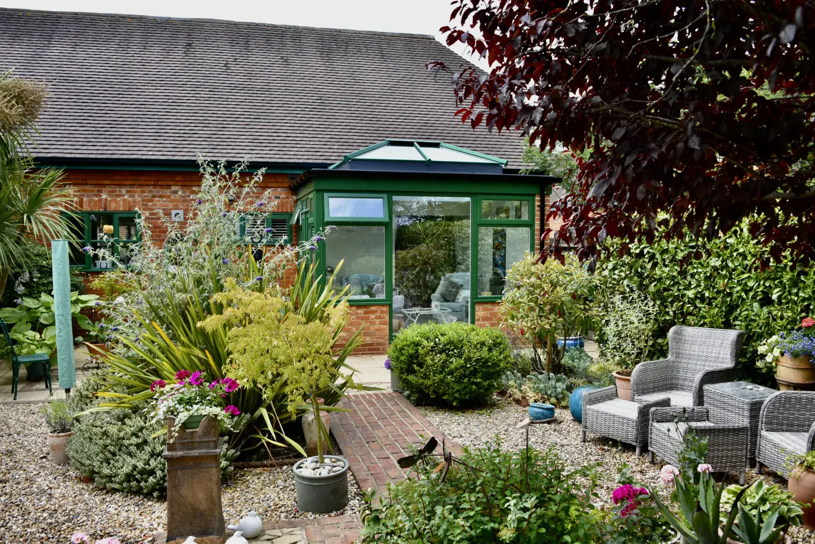 Green framed orangery set behind a planted garden patio with wicker chairs and gravel paths.