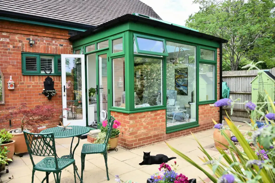 Small green conservatory with brick base, white French doors and patio seating outside.