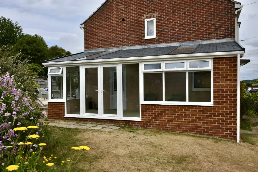 Lean-to conservatory with white frames, tilt windows, French doors and red brick dwarf wall facing a garden.
