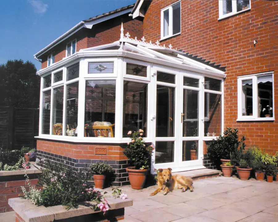 White Victorian conservatory with decorative roof cresting, brick base, patio plants and a dog outside.