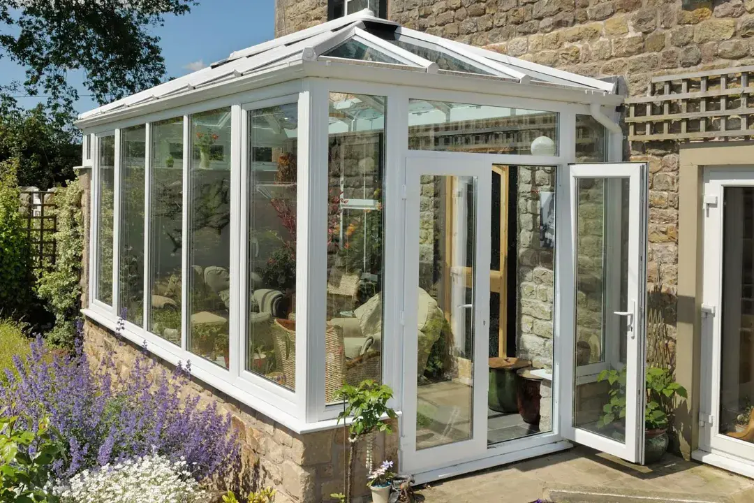 White glass roof conservatory attached to a stone house, with an open door and lavender planting outside.