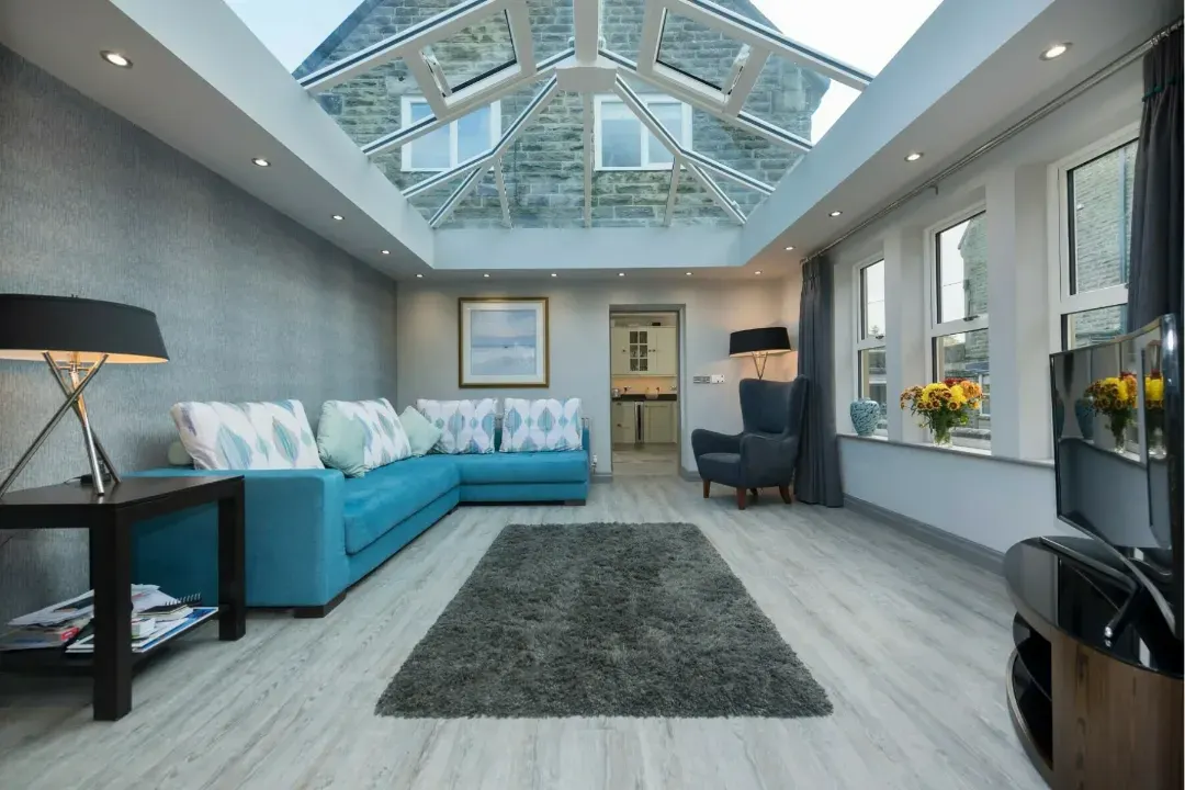 Modern living room with a glass conservatory roof, blue sofa, grey rug and white-framed windows.