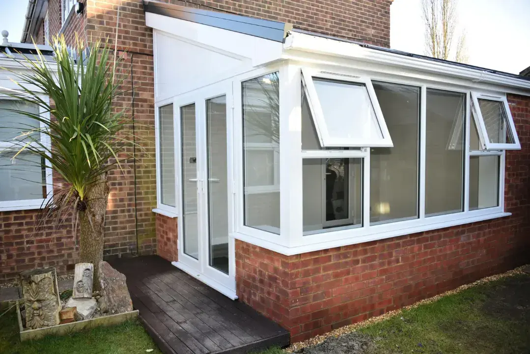 White lean-to conservatory with brick base, side French doors, open top windows and timber decking.