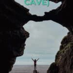 Person standing on a rock in front of the sea, framed by a large cave opening with cloudy sky.
