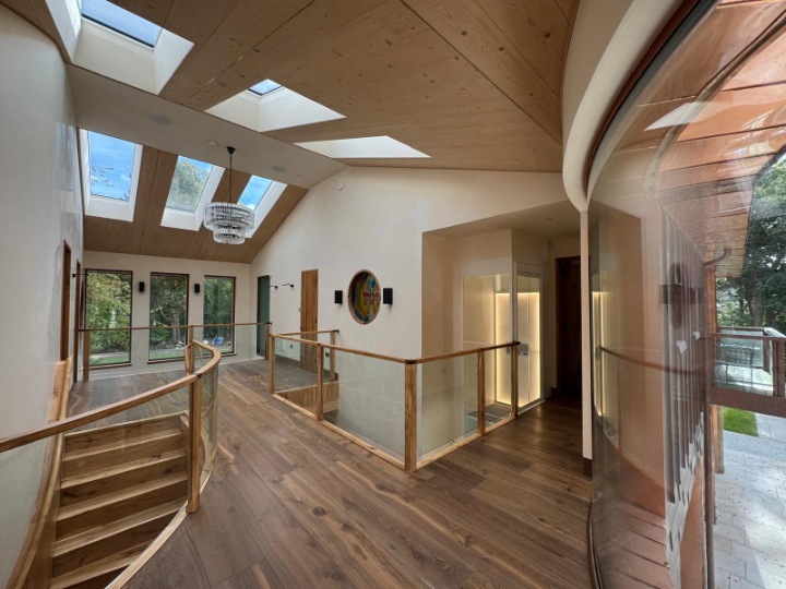 Bright interior hallway with wood flooring, glass balustrades, an glass residential elevator, skylights, and a curved glass wall looking outwards.