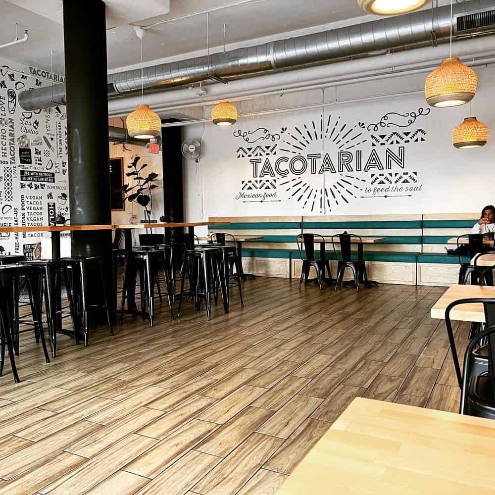 The interior of Tacotarian, a vegan restaurant in North Park, features a modern design with wood-look tile flooring, black metal bar stools, and a large wall mural with the brand name.