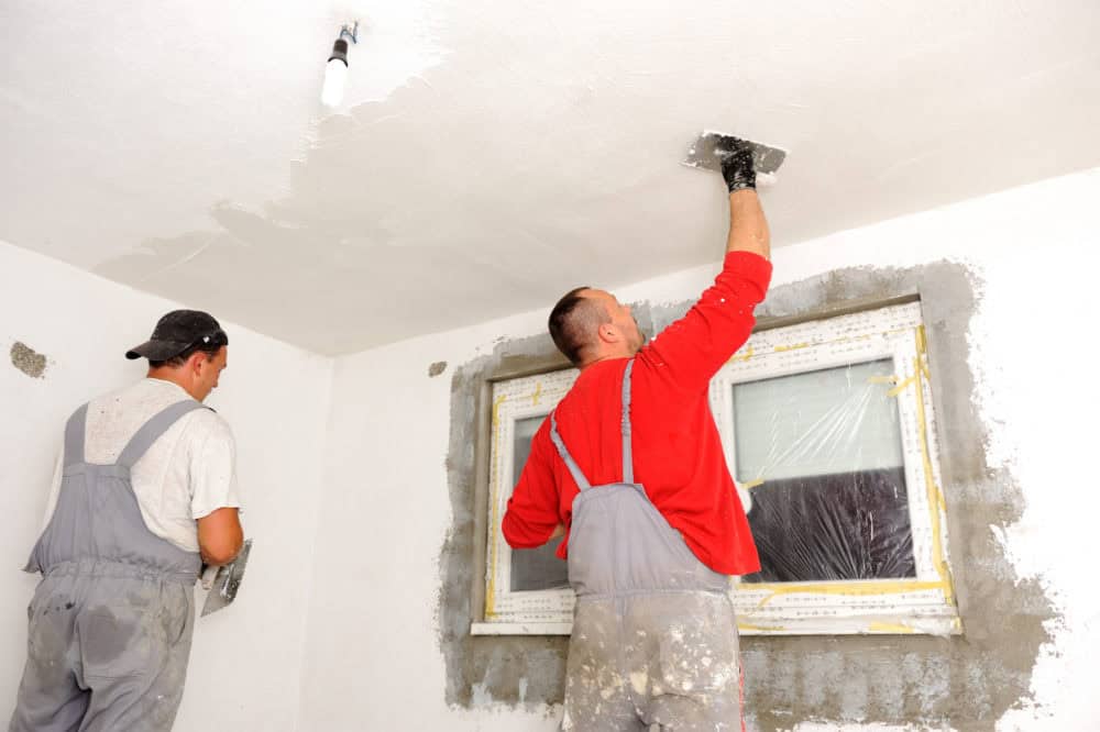 Two construction workers in overalls plastering a white ceiling and walls inside a room under renovation.