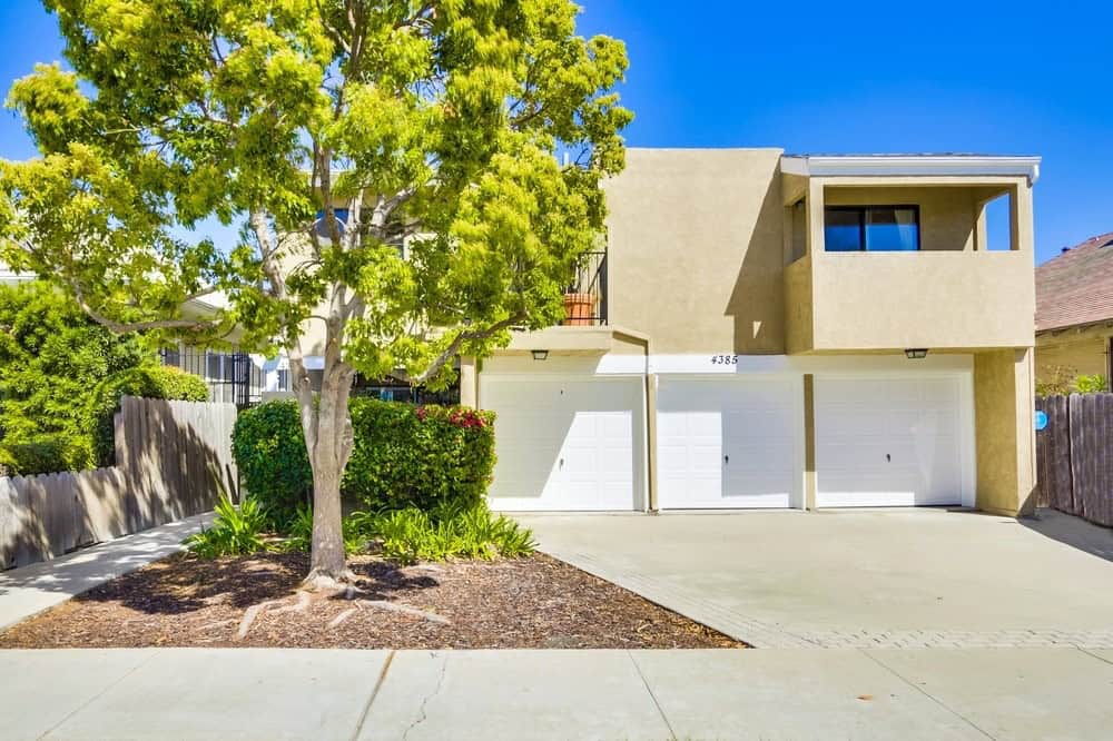 Modern tan multi-unit building at 4385 Georgia Street in University Heights featuring three white garage doors, an upper-level balcony, and a large leafy tree in the front yard.
