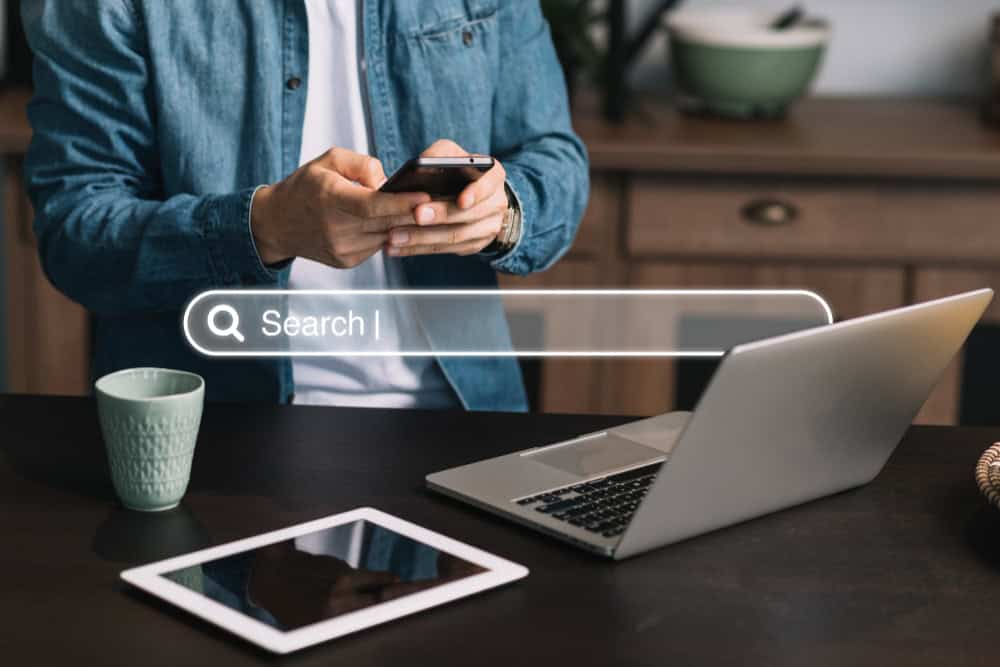 Uma pessoa em camisa jeans usa um smartphone, com uma barra de pesquisa Search flutuando acima de um laptop, tablet e caneca na mesa.